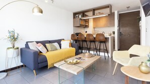 A modern living room with a sofa, coffee table, and chairs, a kitchen area with stools, and a dining table. - Modern Family Apartment in Las Condes (Las Condes)
