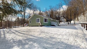 Una casa verde con un porche cubierto, rodeada de nieve en el suelo y entre los árboles.