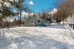 A green house with a covered porch, surrounded by snow-covered ground and trees. - Walk-to-town house, one-mile from Okemo Resort, a/c, w/d, wifi, yard, deck (Ludlow)