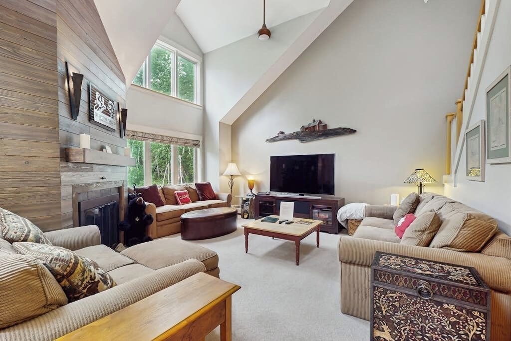 A spacious living room with a high ceiling, a fireplace, a television, and a large window.