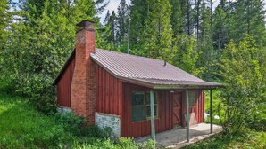 Eine kleine, rote Holzhütte mit Schornstein, umgeben von dichtem Grün und Bäumen.