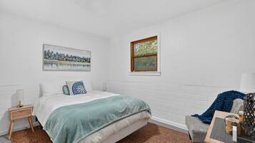 Um quarto com cama, janela, quadro, mesa de cabeceira e uma cadeira.