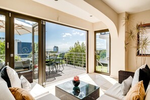 A modern living room with a glass coffee table, a sofa, and a view of a balcony with outdoor furniture. - Homely Sea Point 3bd Villa w Garden Pool Kaya Eden (Cape Town)