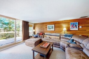 Ein Wohnzimmer mit einer Eckcouch, einem Couchtisch und einem großen Fenster mit Blick ins Freie.