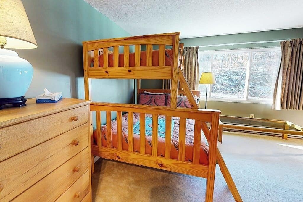 Een stapelbed met houten frame, een nachtkastje met lamp en een raam met gordijnen.