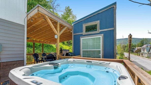 Ein Whirlpool auf einer Holzdiele, eine überdachte Außenlounge und ein blaues Gebäude mit einem großen Fenster.