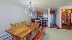 A dining area with a wooden table and chairs, a modern chandelier, and a kitchen in the background.
