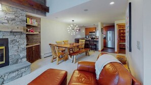 A spacious living area with a stone fireplace, wooden dining table, leather sofa, and a modern kitchen in the background.