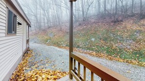 A covered porch with a wooden railing, a house with siding, and a forested area with fallen leaves.