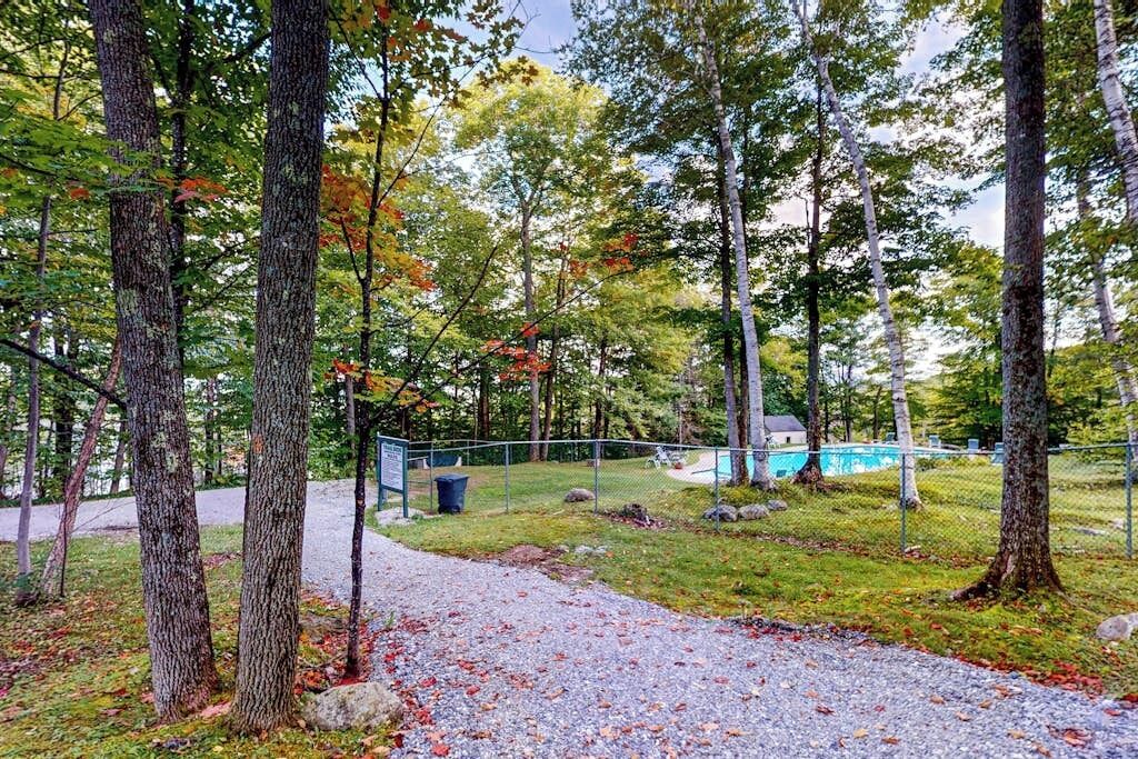 A forested area with a gravel path, a fenced pool area, and a building in the background.