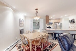 A dining area with a wooden table and chairs, a black leather chair, a chandelier, and a kitchen in the background.