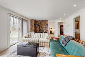 A modern living room with a sofa, ottoman, and a green couch. There is a brick wall and a fireplace. - Three-story Ludlow Mountain home with private hot tub, firepit and EV charger (Ludlow)
