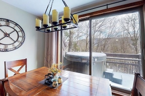 A dining area with a wooden table, chairs, a wall clock, and a window with a view of a hot tub and trees. - Rustic home with a private hot tub, cable, & washer/dryer (Plymouth)