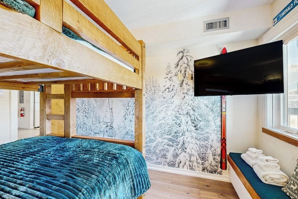 Une chambre avec un lit superposé, un téléviseur fixé au mur, une fenêtre donnant sur des arbres couverts de neige et un banc avec des serviettes.