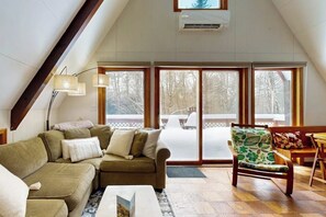 Ein gemütliches Wohnzimmer mit einer Couch, einem Sessel und einem Tisch. Große Fenster bieten Blick auf Bäume.