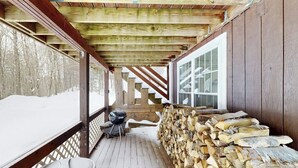 A covered wooden deck with a grill, stacked firewood, and a view of a snowy landscape. - Cozy duplex, first floor unit w/ kitchen, shared fire pit, lake access, wifi, TV (Ludlow)