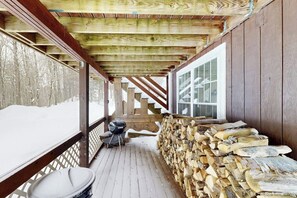 A covered wooden deck with a grill, stacked firewood, and a view of a snowy landscape. - Cozy duplex, first floor unit w/ kitchen, shared fire pit, lake access, wifi, TV (Ludlow)
