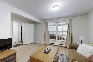 A modern living room with a television, a coffee table, a sofa, and a large window with curtains. - Cozy duplex, first floor unit w/ kitchen, shared fire pit, lake access, wifi, TV (Ludlow)