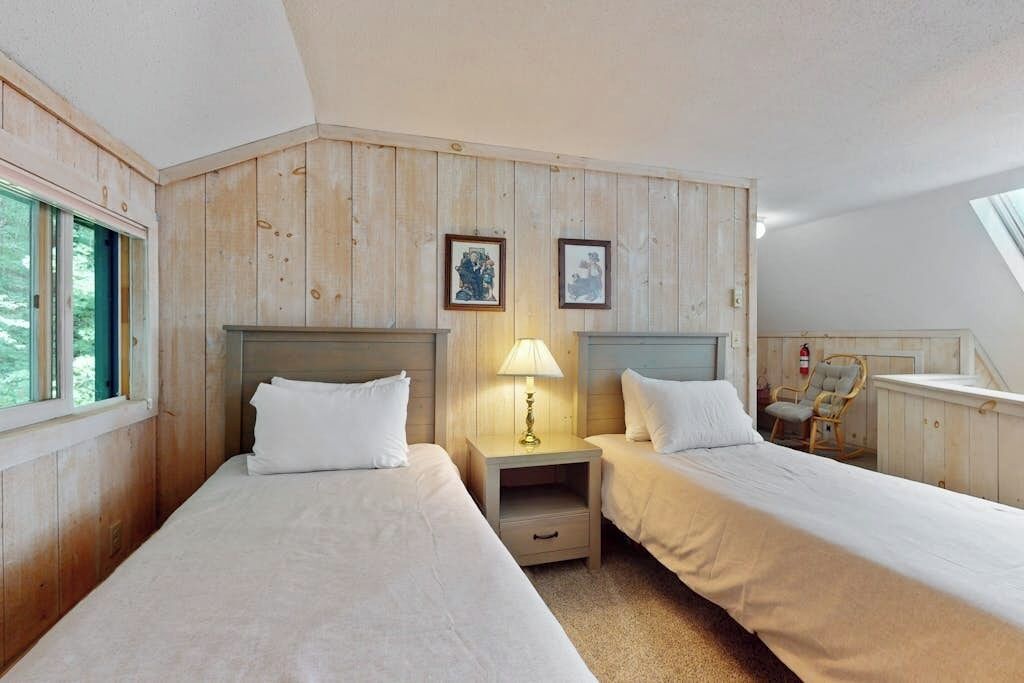 Una habitación con dos camas, paneles de madera, una mesita de noche con lámpara, cuadros enmarcados en la pared y una silla.