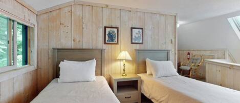 Una habitación con dos camas, paneles de madera, una mesita de noche con lámpara, cuadros enmarcados en la pared y una silla.