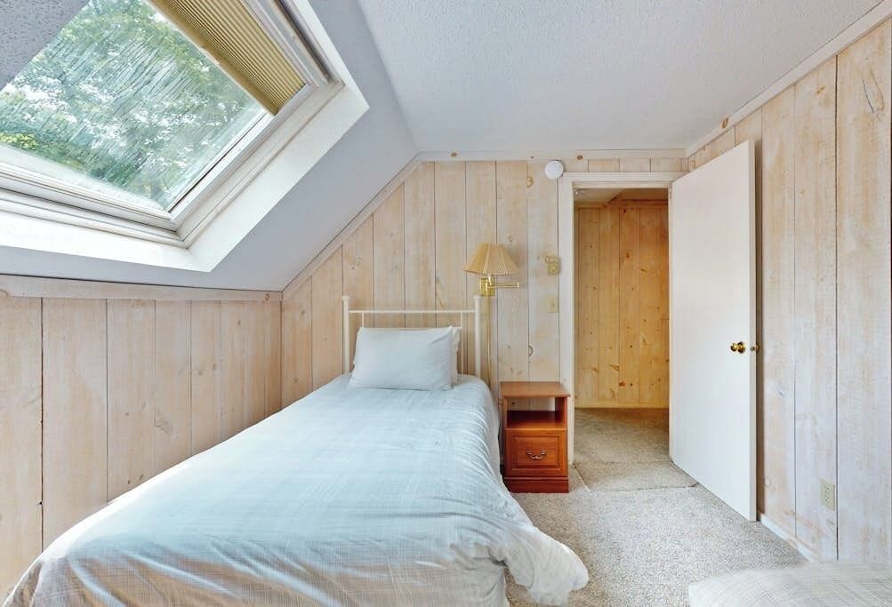 A bedroom with a skylight, a bed, a bedside table with a lamp, and a door leading to another room.