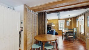 A rustic interior with a wooden dining table, blue bar stools, and a kitchen area visible through an open door.
