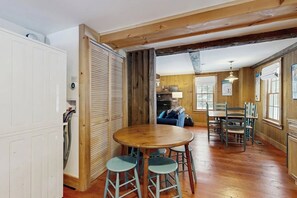 A rustic interior with a wooden dining table, blue bar stools, and a kitchen area visible through an open door.