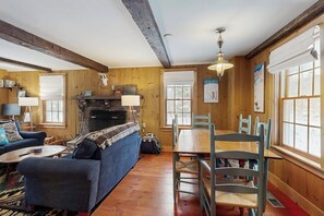 A cozy living room with a fireplace, wooden walls, a sofa, a dining table with chairs, and a large window.