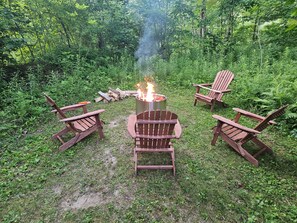 A campfire with wooden chairs arranged around it in a forest setting.