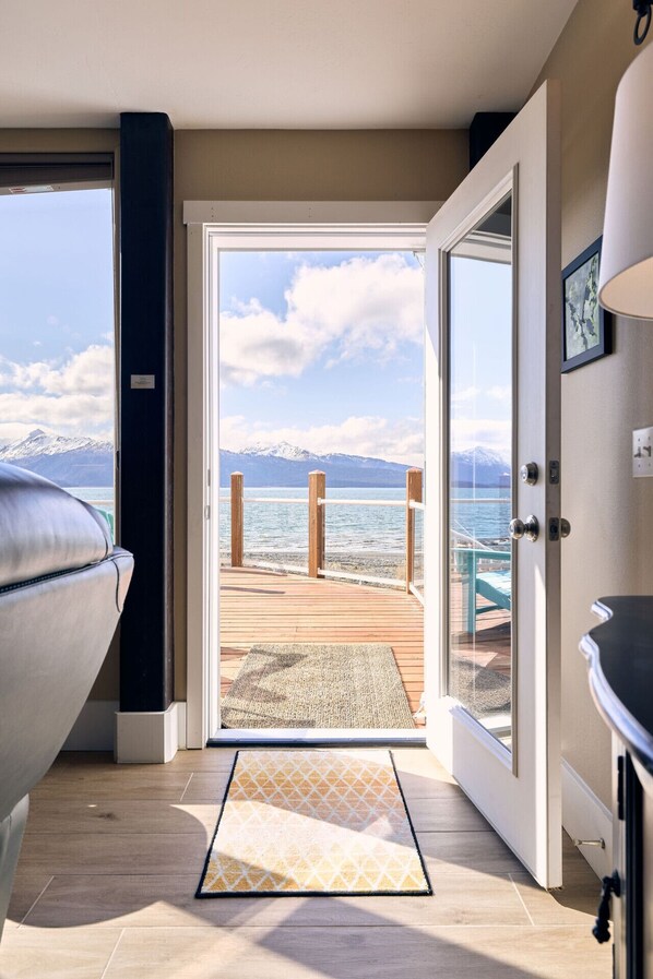 A room with a view of a beach and mountains, a wooden deck, and a grill.