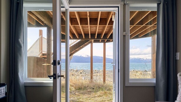 Ein Zimmer mit einer großen Schiebeglas-Tür, die auf einen Balkon mit Holzgeländer und Blick auf Strand und Berge führt.