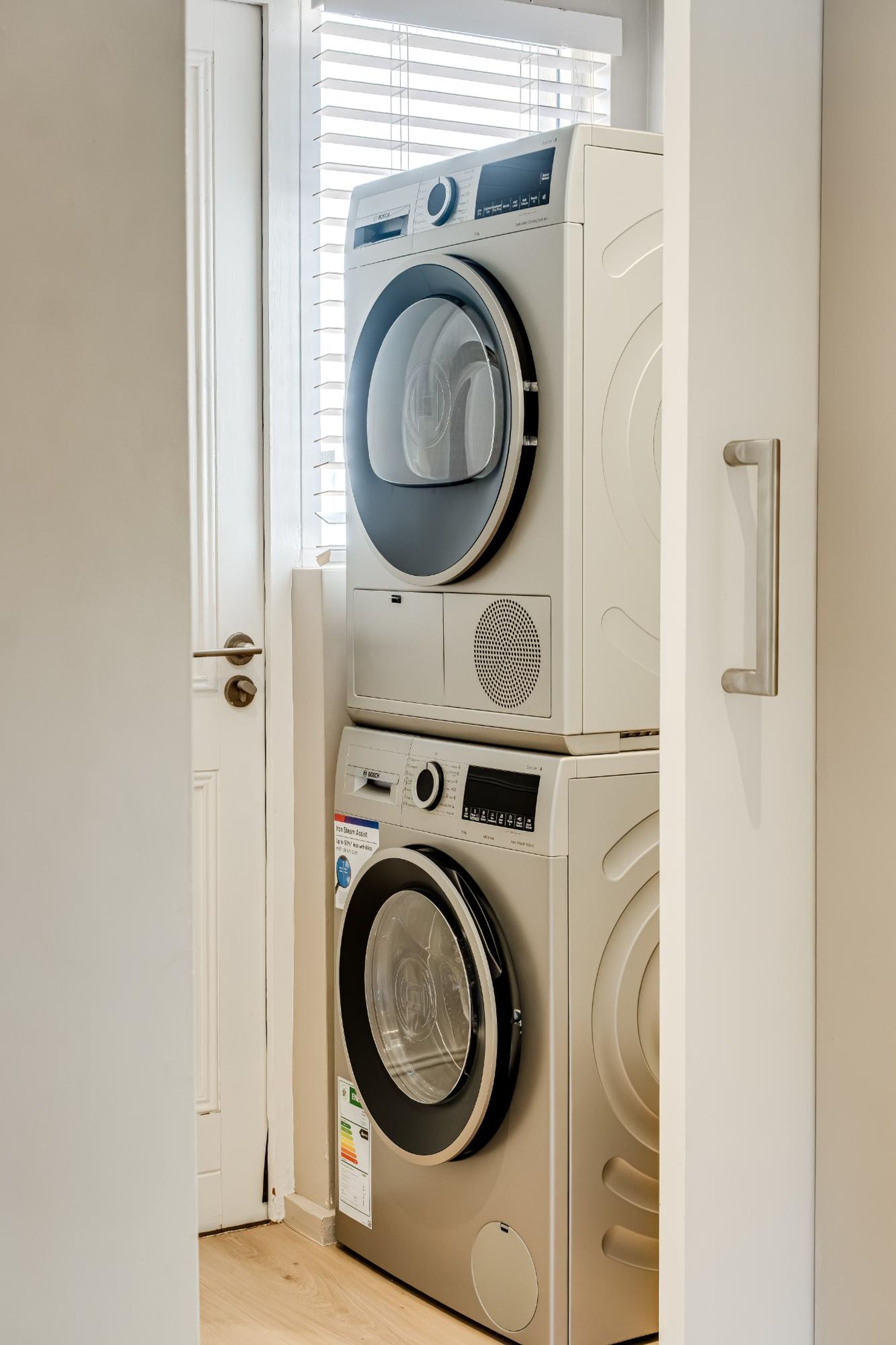A laundry room with stacked washing machines and dryers, a door, and a window with blinds.
