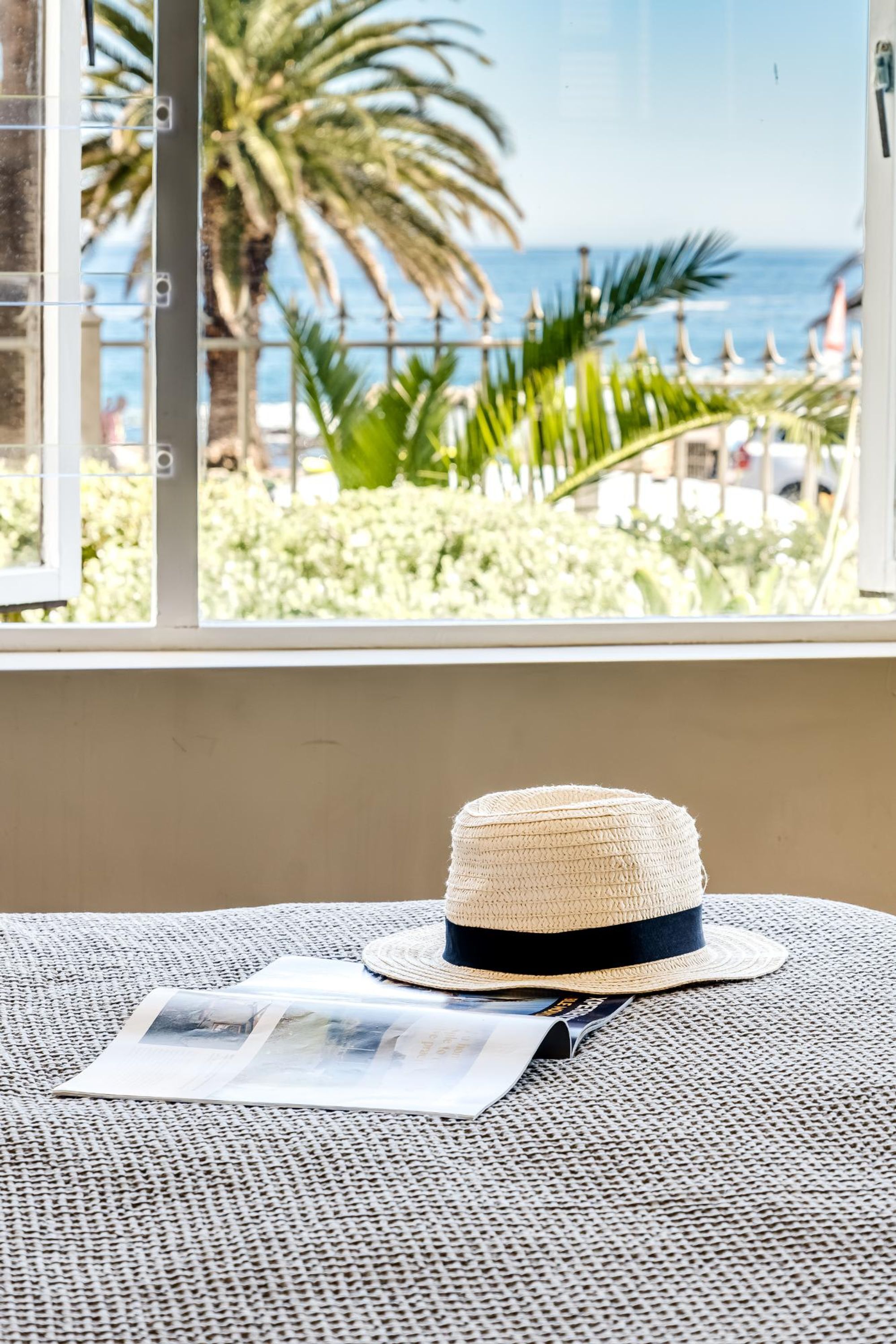 A straw hat on a woven mat, a magazine, and a window with a view of palm trees and the sea.