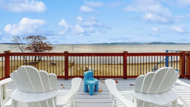 A deck with white chairs and a blue stool overlooking a beach.