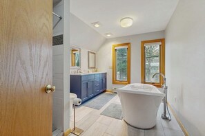 A modern bathroom with a freestanding bathtub, a vanity with a mirror, and two windows.