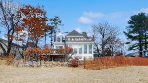 A coastal house with a large bay window, a deck, and a sandy beach in the foreground.