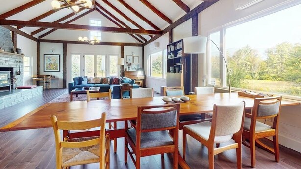 Amplia sala de estar con una mesa de madera para comer, sillas, chimenea y un ventanal con vistas a la vegetación.