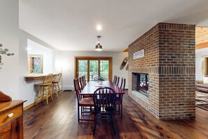 A dining area with a brick fireplace, wooden flooring, and a large window.