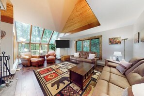 A spacious living room with a high ceiling, large windows, a flat-screen TV, leather sofas, and a patterned rug.