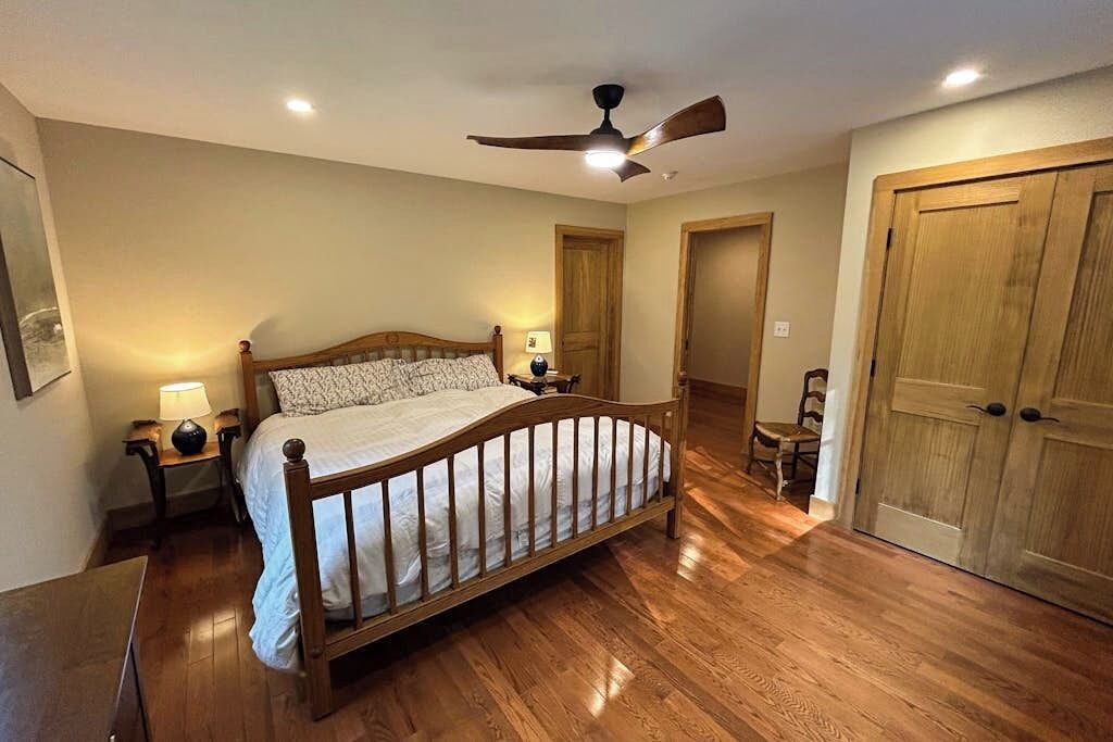 Een slaapkamer met een houten bed, een plafondventilator, een nachtkastje met een lamp en een houten deur.