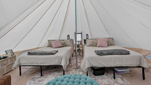 Uma tenda com duas camas, cada uma com almofadas e cobertores, uma mesinha e um banquinho azul.