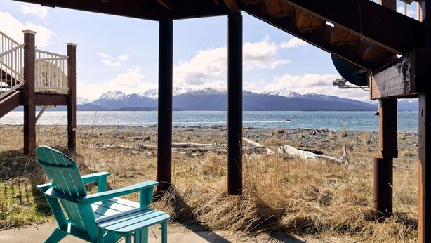 Un espacio de estar al aire libre cubierto, con sillas, una terraza de madera y vista a un lago con montañas al fondo.