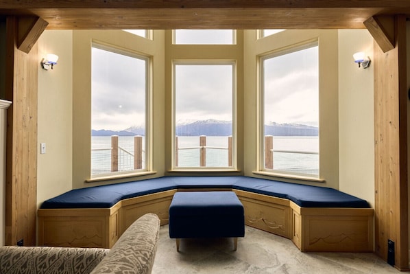 A room with a bay window seating area, wooden walls, and a view of a body of water with mountains in the distance.