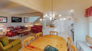 A spacious living area with a wooden dining table, two red and green sofas, a green armchair, and a kitchen in the background.