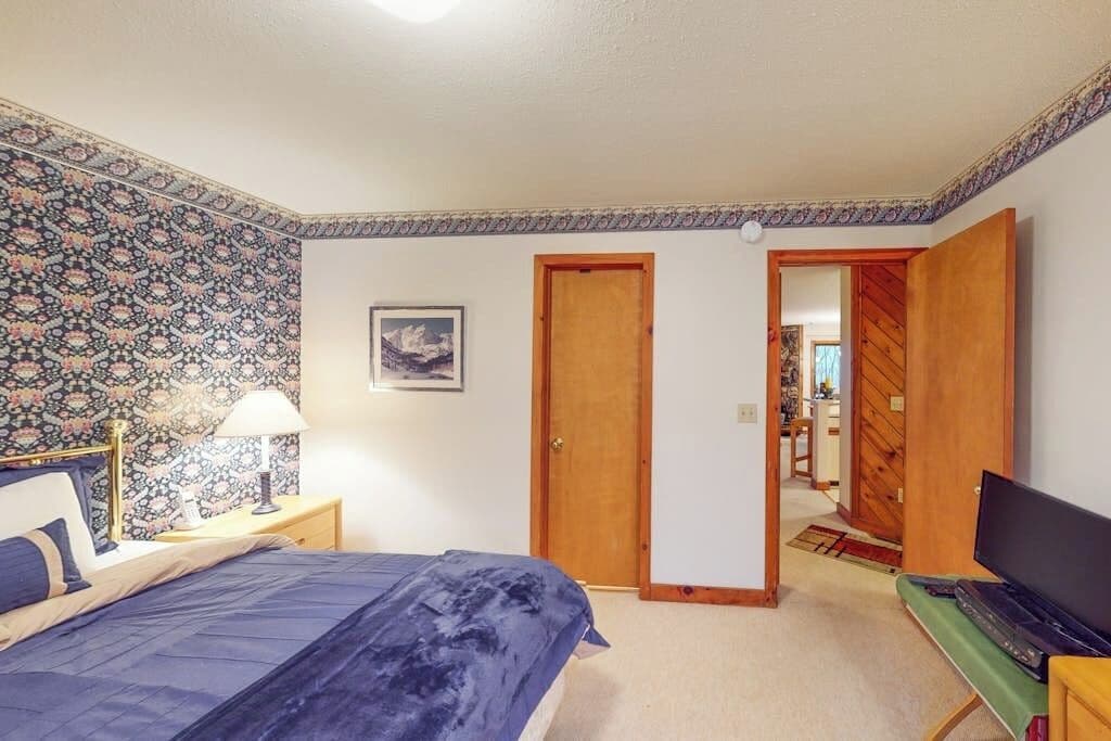 A bedroom with a bed, a nightstand, a lamp, a television, and a door leading to another room.