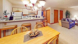 A dining area with a wooden table, chairs, and a chandelier. There is a kitchen in the background with a refrigerator and microwave.