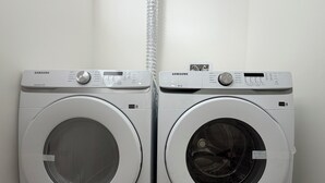 Two white Samsung front-loading washing machines in a laundry room with a white wall and a metal vent. - Brand new basement suite with private entrance, 3-5 minutes to market and parks (Saskatoon)