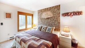 Een slaapkamer met een bed, een nachtkastje, een ventilator aan de muur, een raam en een lijst met een afbeelding.
