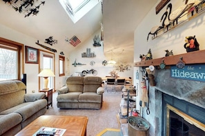 A cozy living room with a fireplace, a sofa, a coffee table, and a dining area in the background. - Comfortable Townhome with Shared Pool, Private Hot Tub & Wood-Burning Fireplace (Stratton)