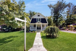 A two-story house with a front porch, a well-maintained lawn, and a pathway leading to the entrance. - Classic village home with large porch, washer/dryer, cable, grill & partial AC (Manchester)
